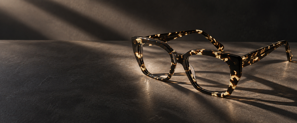 Framed eyeglasses with a tortoiseshell pattern in black and gold, resting on a dark textured surface under dramatic diagonal light beams.